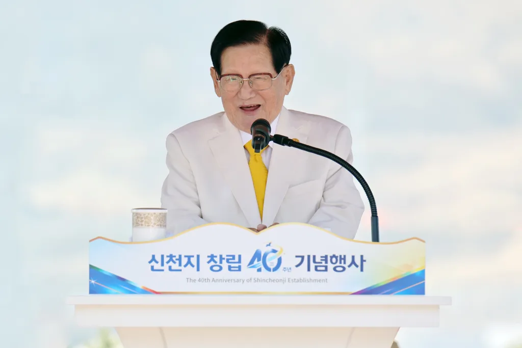 Pohled na předsedu církve, pana Lee Man Hee, který má slovo na oslavě výročí církve Shincheonji. Má na sobě bílý oblek a žlutou kravatu, usmívá se a mluví do mikrofonu.